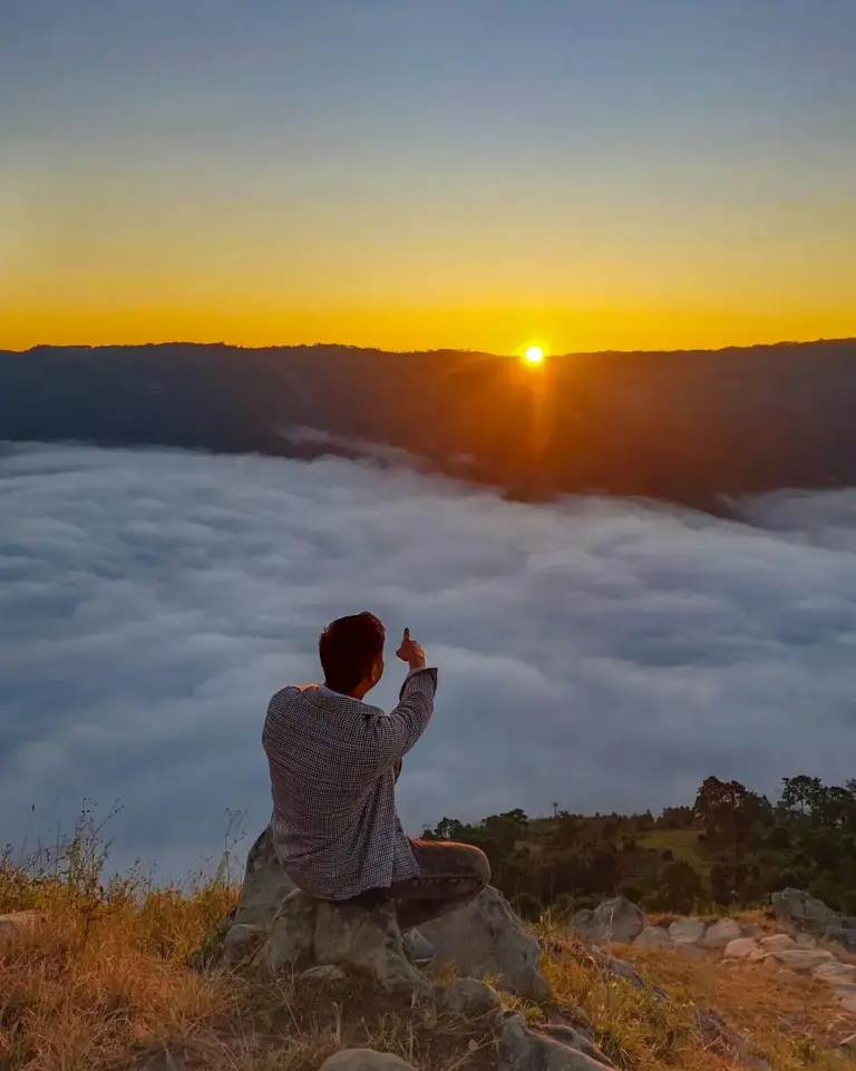 A man sitting on a rock captures the sunrise over a sea of clouds in Nongjrong, Meghalaya