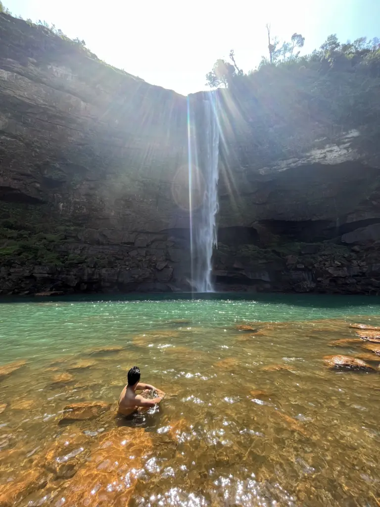 A person relaxing in the shallow, clear waters beneath Phe Phe Falls in Meghalaya, with sunlight streaming down over a tall, narrow waterfall cascading from a rocky cliff