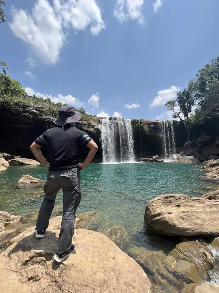 A person standing on a rock, admiring the turquoise blue pool and cascading waters of Krang Suri Falls in Meghalaya under a bright blue sky