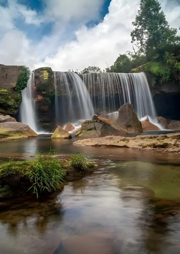 Janailar Falls in Meghalaya, graceful waterfall cascading over rocky cliffs into a clear, shallow pool surrounded by mossy boulders and greenery