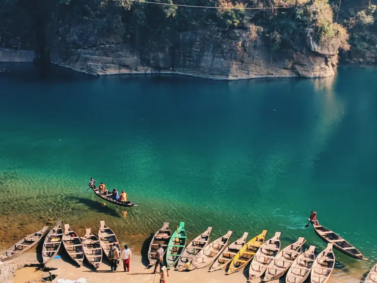 People boarding wooden boats along the crystal-clear waters of the Umngot River in Dawki, with steep rocky cliffs and lush greenery in the background