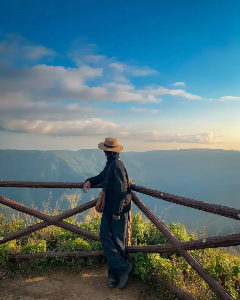Person in a hat leaning on a wooden fence, enjoying the mountain view at Laitlum, Meghalaya
