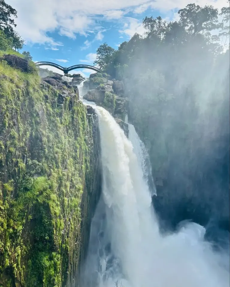 Powerful stream of Weinia Falls cascading down rocky cliffs in Meghalaya, with a misty spray and a scenic bridge above
