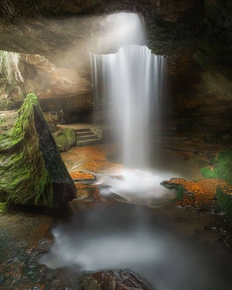 Small waterfall flowing through a rock opening into a mossy cave at the Garden of Caves, Meghalaya