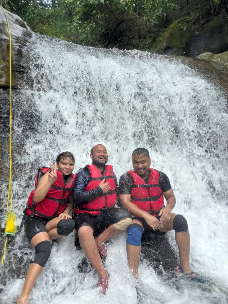 Three people in life jackets sitting under a waterfall at Mawlyngbna Meghalaya