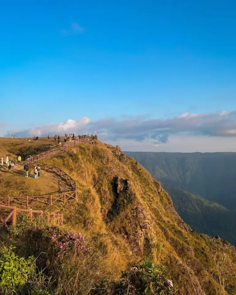 Tourists walking along a fenced path on a grassy cliff at Laitlum, Meghalaya, with a deep valley in the background