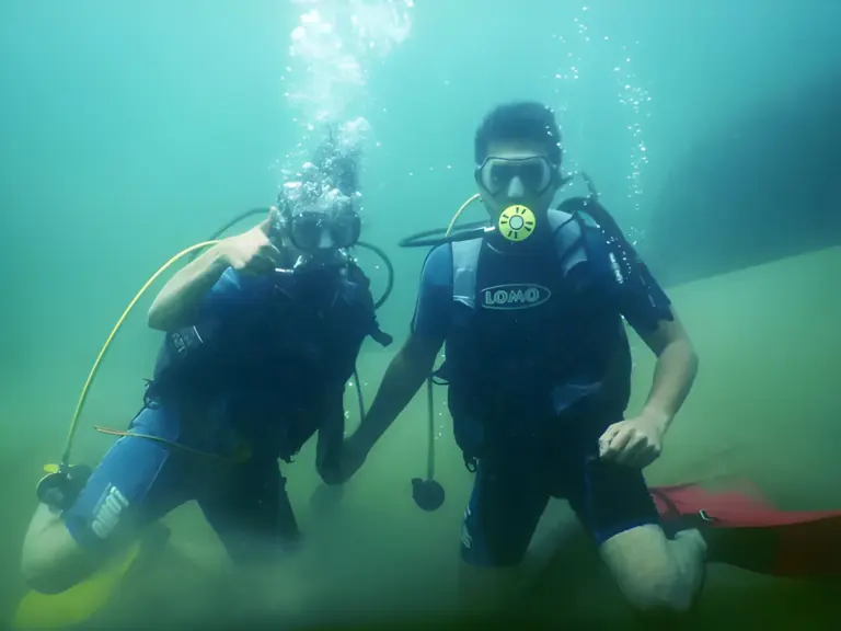 Two people scuba diving underwater in Dawki, wearing Swimsuits and breathing equipment, surrounded by greenish-clear river water