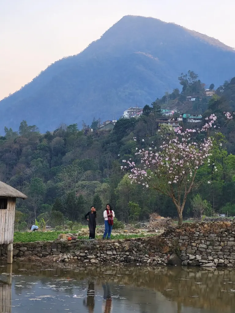 Two women standing by a pond with blooming tree and hills in Dzukou Valley, Nagaland