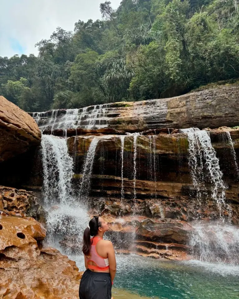 Woman admiring a tiered waterfall surrounded by rocky cliffs and lush forest at Nohkalikai Falls, Meghalaya