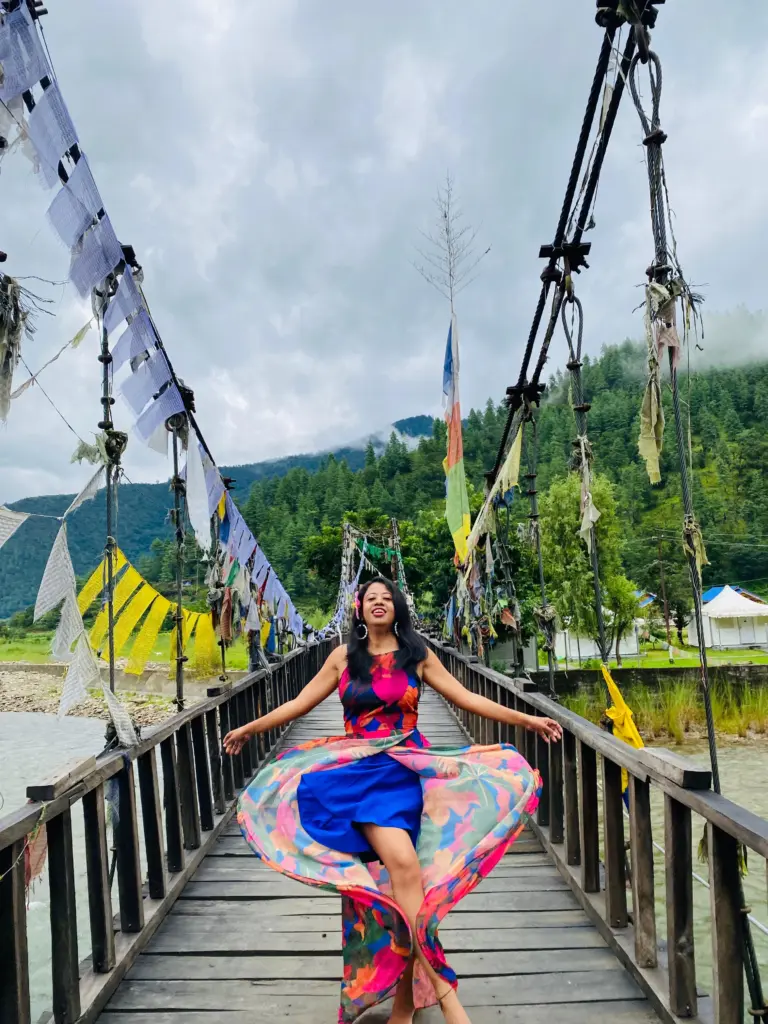 Woman in a colorful dress twirling on a prayer flag-decorated bridge at Sangti Valley