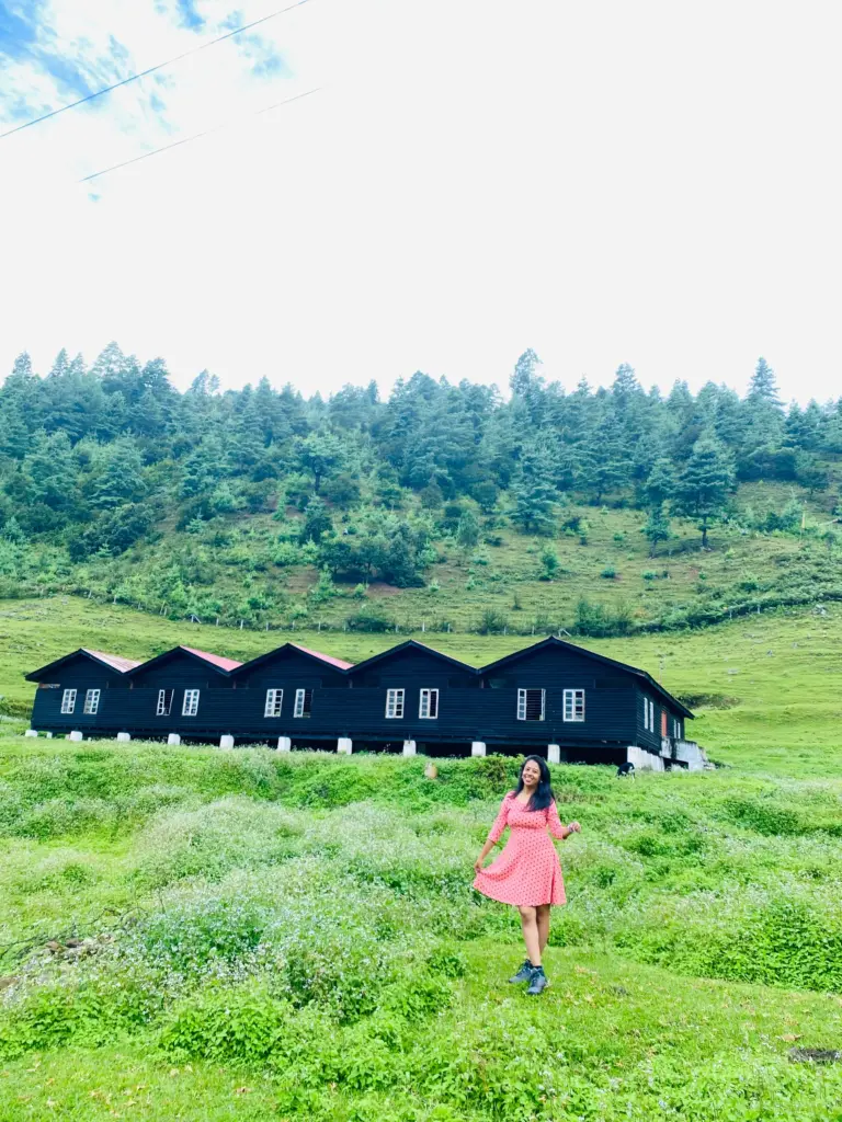 Woman in a red dress standing on green field with wooden cottages and pine trees at Sangti Valley