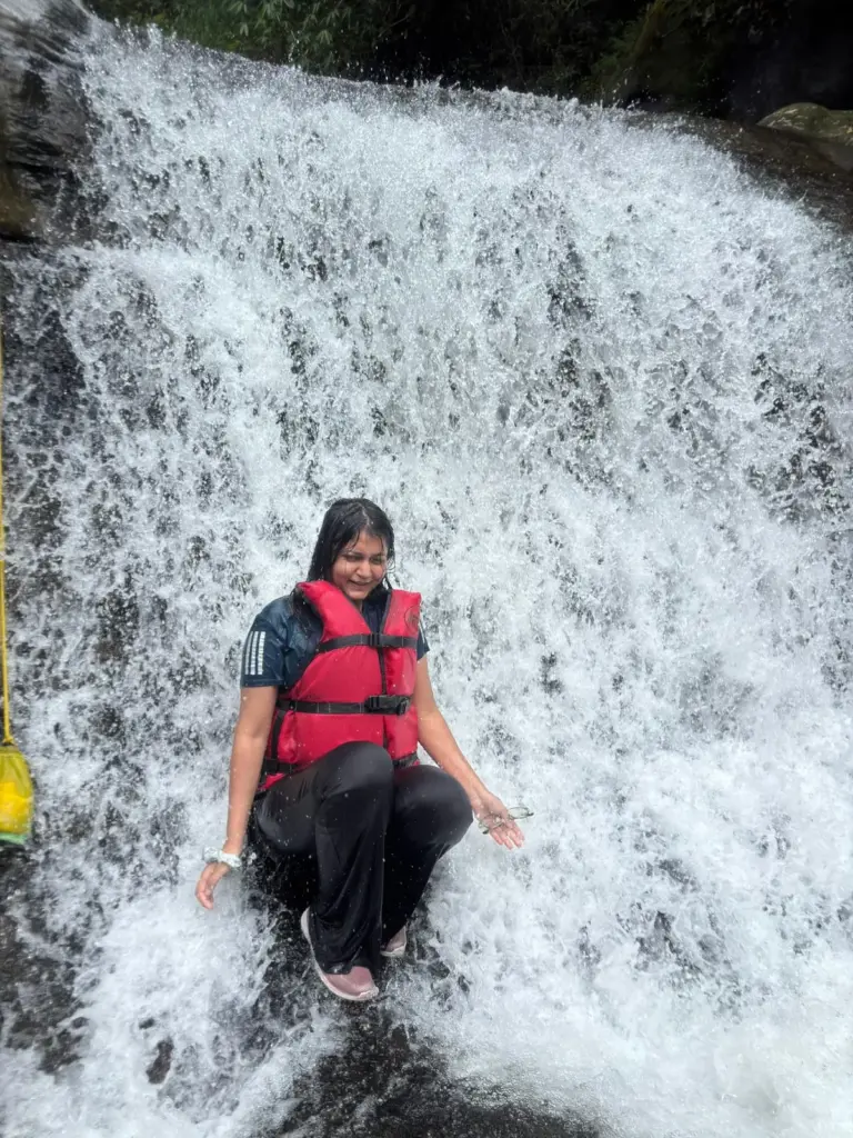 Woman sitting under a waterfall wearing a red life jacket during river canyoning at Mawlyngbna, Meghalaya