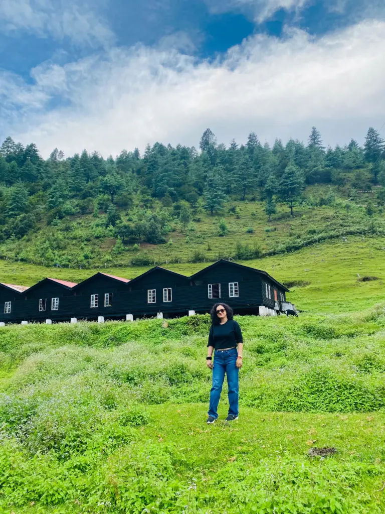 Woman standing on a green meadow with wooden cottages and pine trees at Sangti Valley