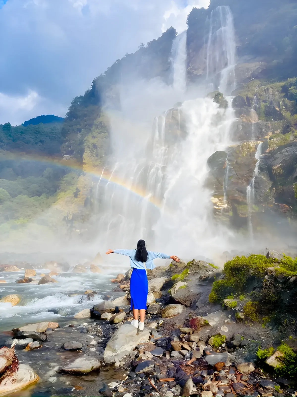 A woman stands on rocks with outstretched arms before Nuranang Falls in Tawang, surrounded by mist and a vibrant rainbow