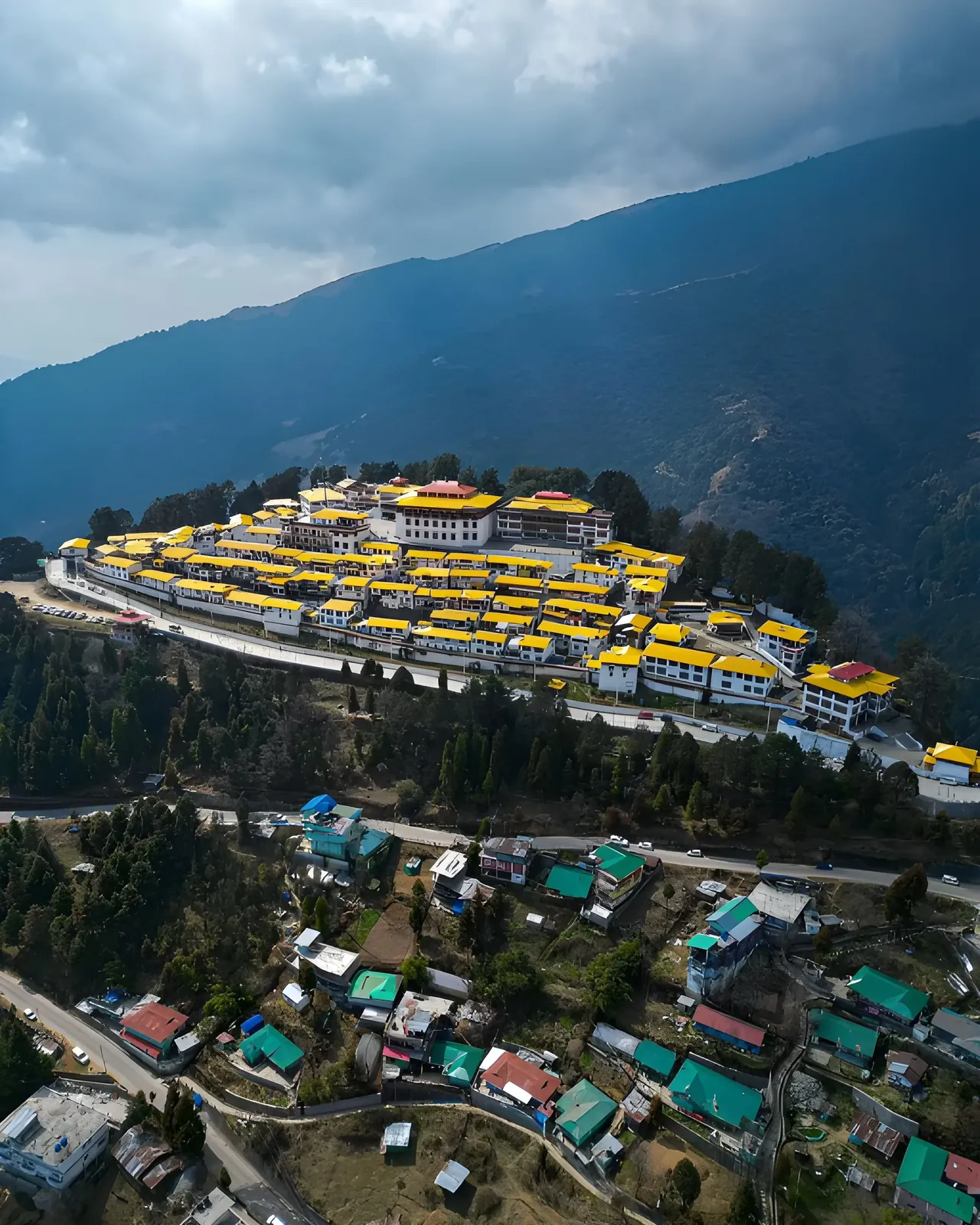 Aerial view of Tawang Monastery with yellow-roofed buildings set against mountain slopes and dramatic clouds