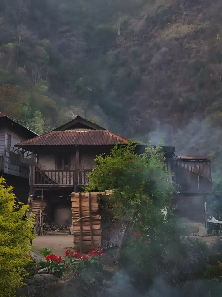 Traditional bamboo houses at the base of a forested hillside, surrounded by flowers and drifting mist